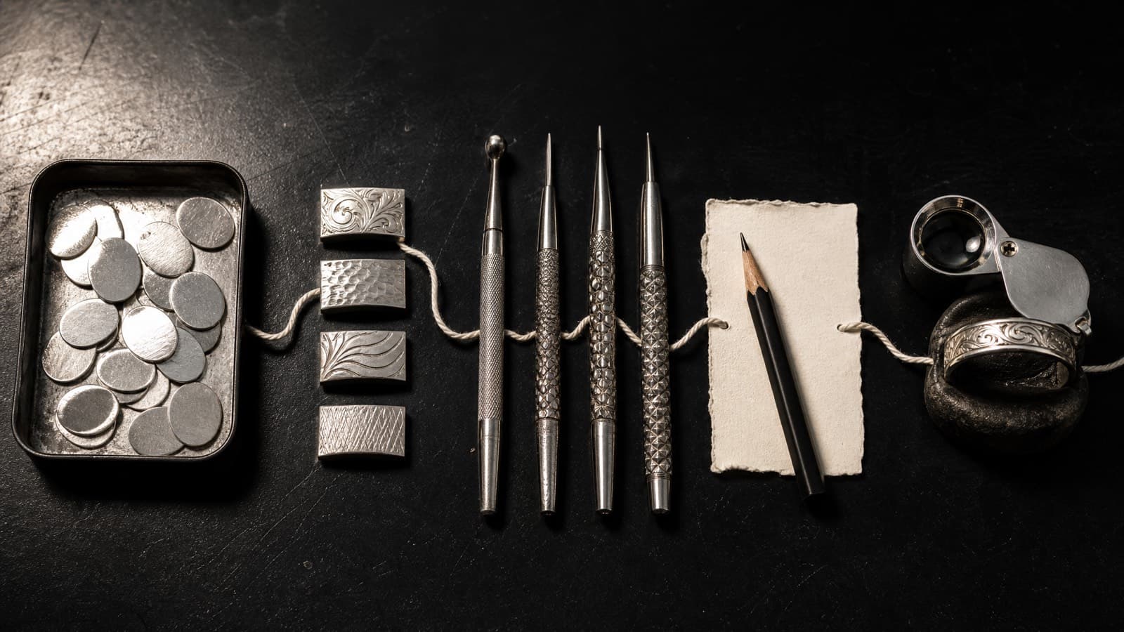 A tray of silver blanks, metal sample coupons, engraving tools and a paper card laid out on a bench