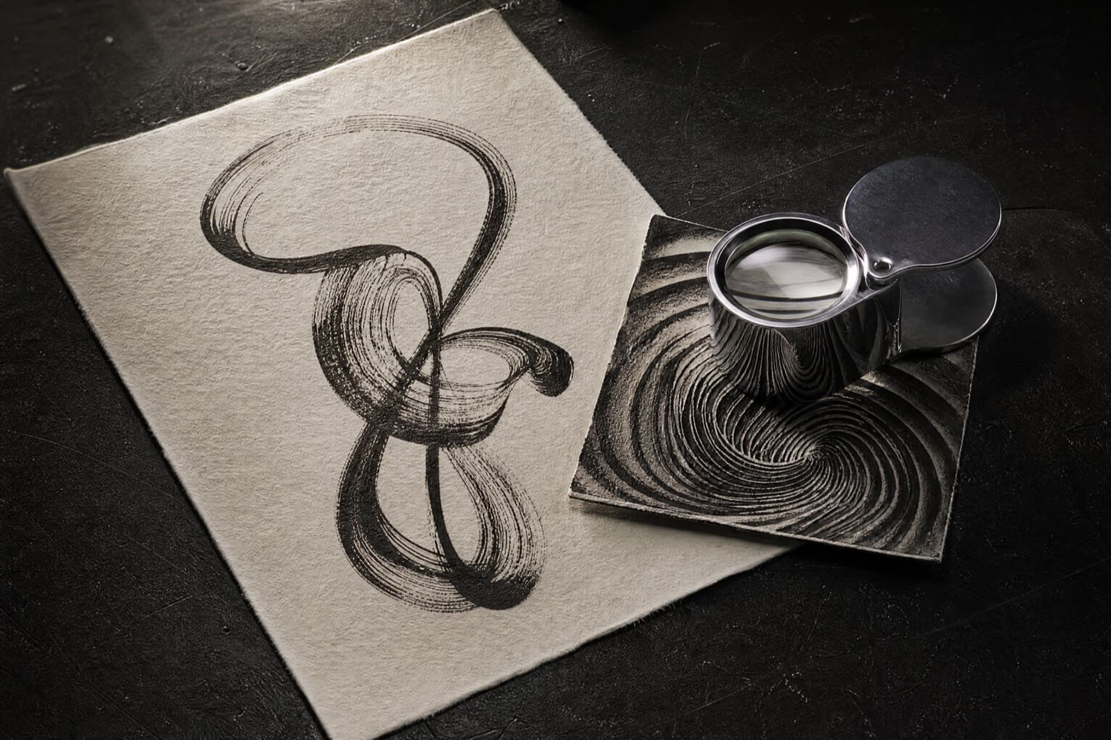 A looping ink stroke on paper, a square of ridged pattern, and a jeweller's loupe on a bench